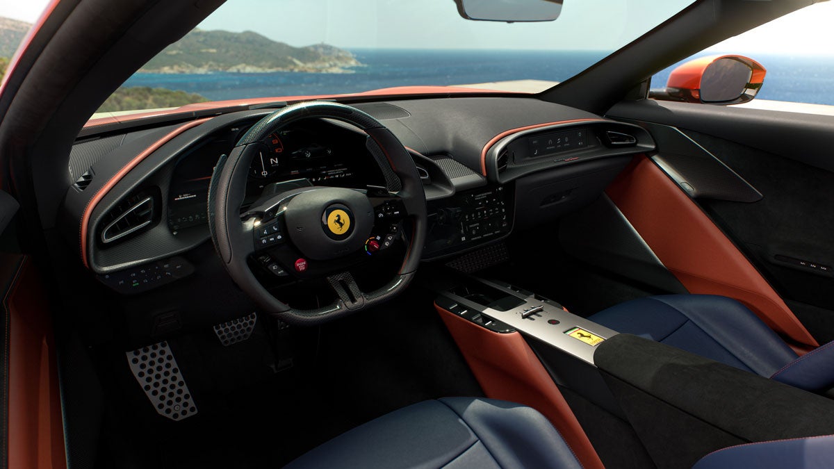Interior view of a modern Ferrari sports car, featuring a black steering wheel with a yellow Ferrari logo, black and orange dashboard, blue seats, and a view of the ocean.