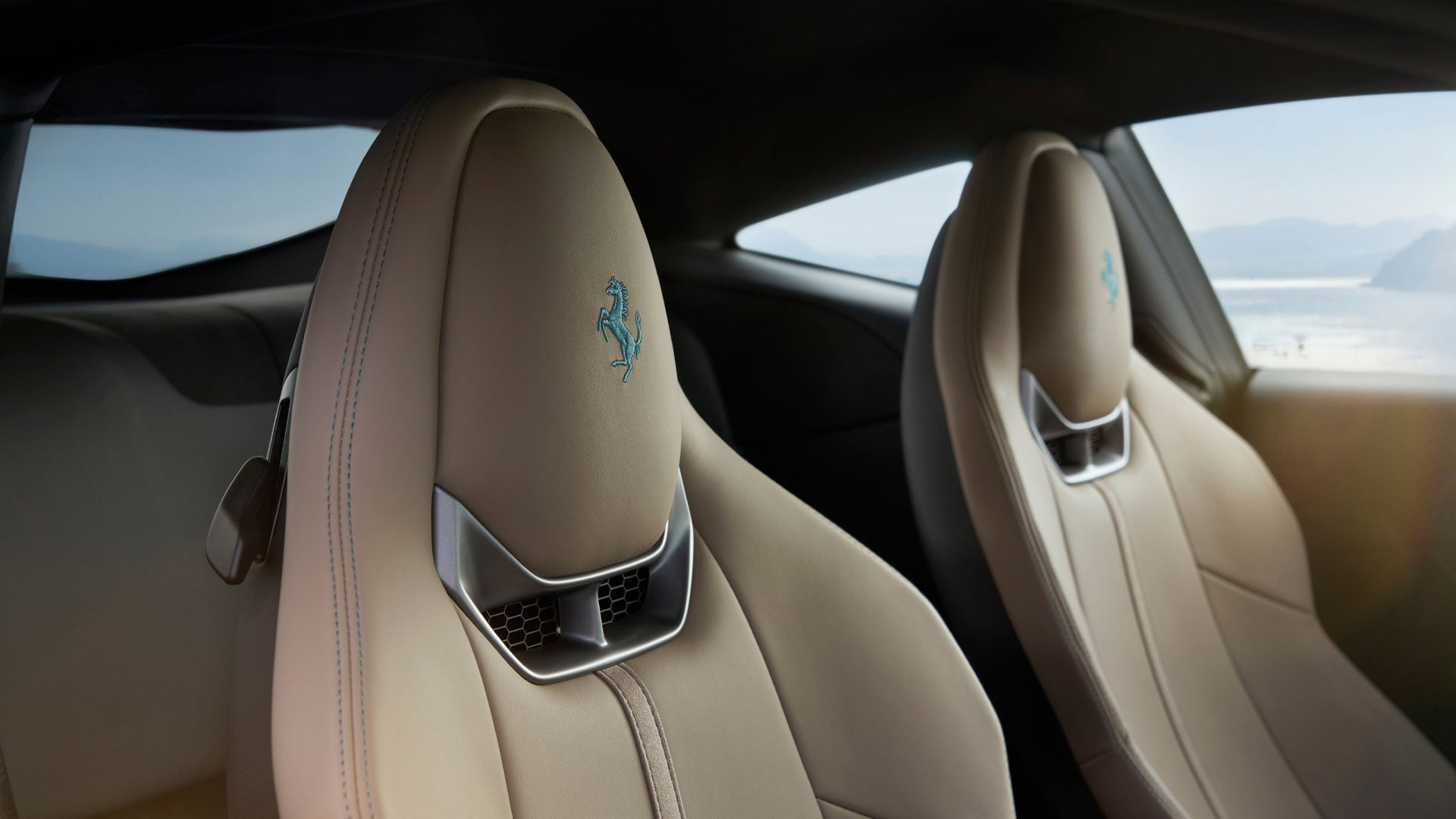 Two beige leather car seats with a blue embroidered Ferrari logo on the headrests, facing forward.