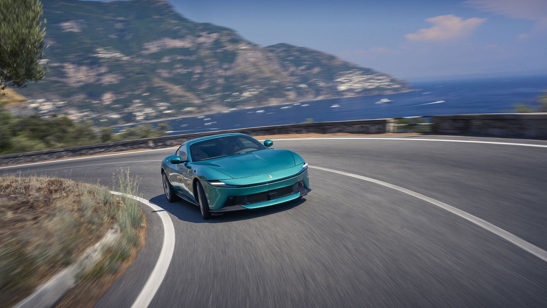 A teal sports car takes a sharp turn on a winding coastal road, with mountains and the sea in the background.