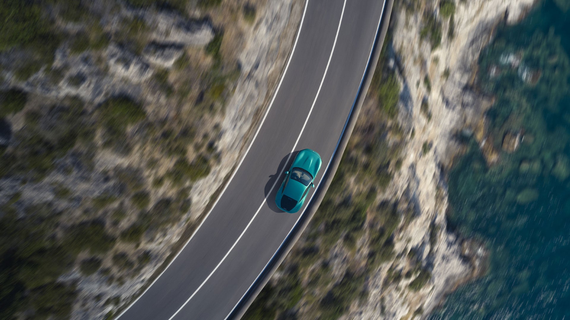 Aerial view of a teal car driving on a coastal road with blur, suggesting motion.
