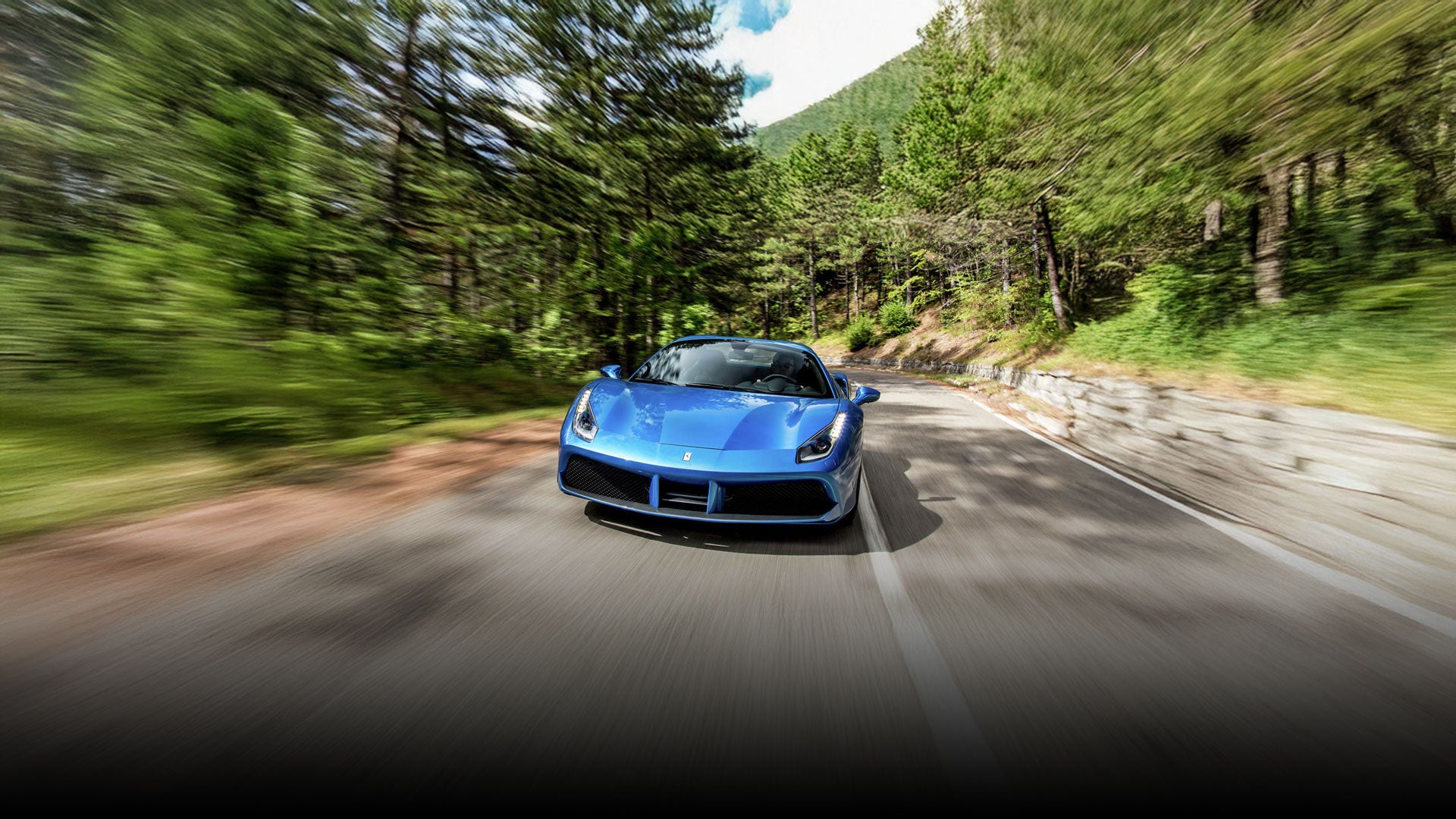 A blue Ferrari sports car speeding down a winding road lined with green trees.